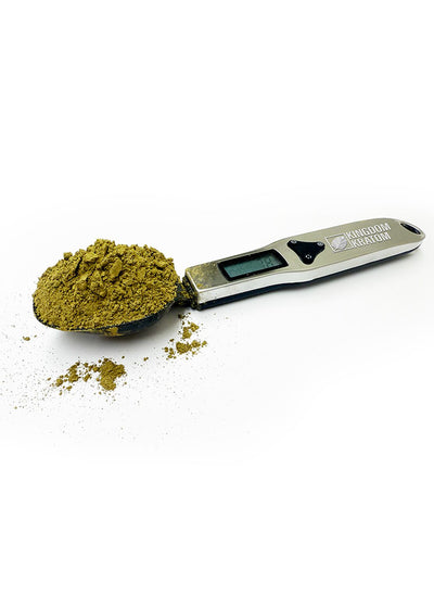 A digital measuring spoon with a metal spoon and ABS plastic body, displaying an LCD screen showing the weight of the substance. Some kratom powder is scattered around the spoon.